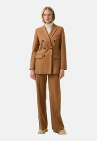 Brązowa marynarka dwurzędowa z dwoma przednimi kieszeniami, noszona na jasnym swetrze z wysokim kołnierzem, w zestawie z dopasowanymi spodniami i jasnobeżowymi butami.