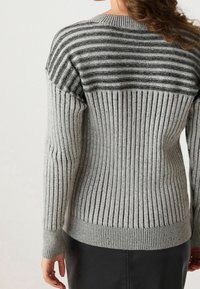 Femme portant un pull en maille côtelée gris avec des rayures horizontales dans le haut du dos et des rayures verticales en dessous, associé à une jupe noire.