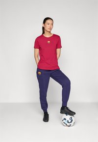 Rød bomull T-skjorte med logo, sammen med marineblå joggebukser med en lignende logo. Svarte treningssko på en fotball.