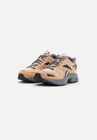 Paire de chaussures de sport Reebok beige et noire avec des semelles et des lacets gris sur un fond blanc uni.