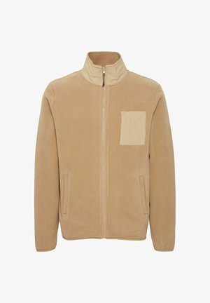 Veste en polaire beige avec un col montant, fermeture éclair à l'avant, poches latérales et une poche poitrine carrée dans une teinte légèrement plus claire. Texture douce.