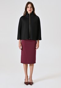 Capa negra con cremallera sobre una falda lápiz burdeos, combinada con tacones puntiagudos burdeos, contra un fondo blanco liso.