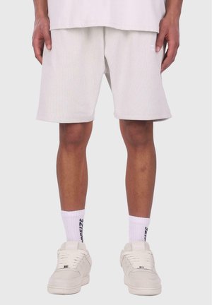 Hvite ribbede shorts med løs passform, midt på låret, kombinert med hvite joggesko og teksturerte hvite sokker med svart tekst.