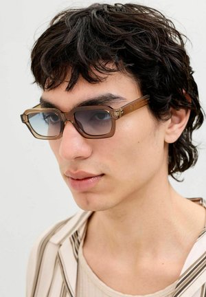 Jauns vīrietis ar īsiem tumšiem cirtainiem matiem, valkājot brūnas taisnstūra formā saulesbrilles un bēšu krāsas svītrainu kreklu, raugās uz kreiso pusi.