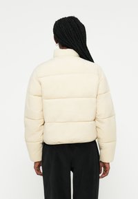 Giacca imbottita color crema con colletto alto, design trapuntato e maniche lunghe, indossata sopra pantaloni neri. Tessuto semplice, senza motivi visibili.