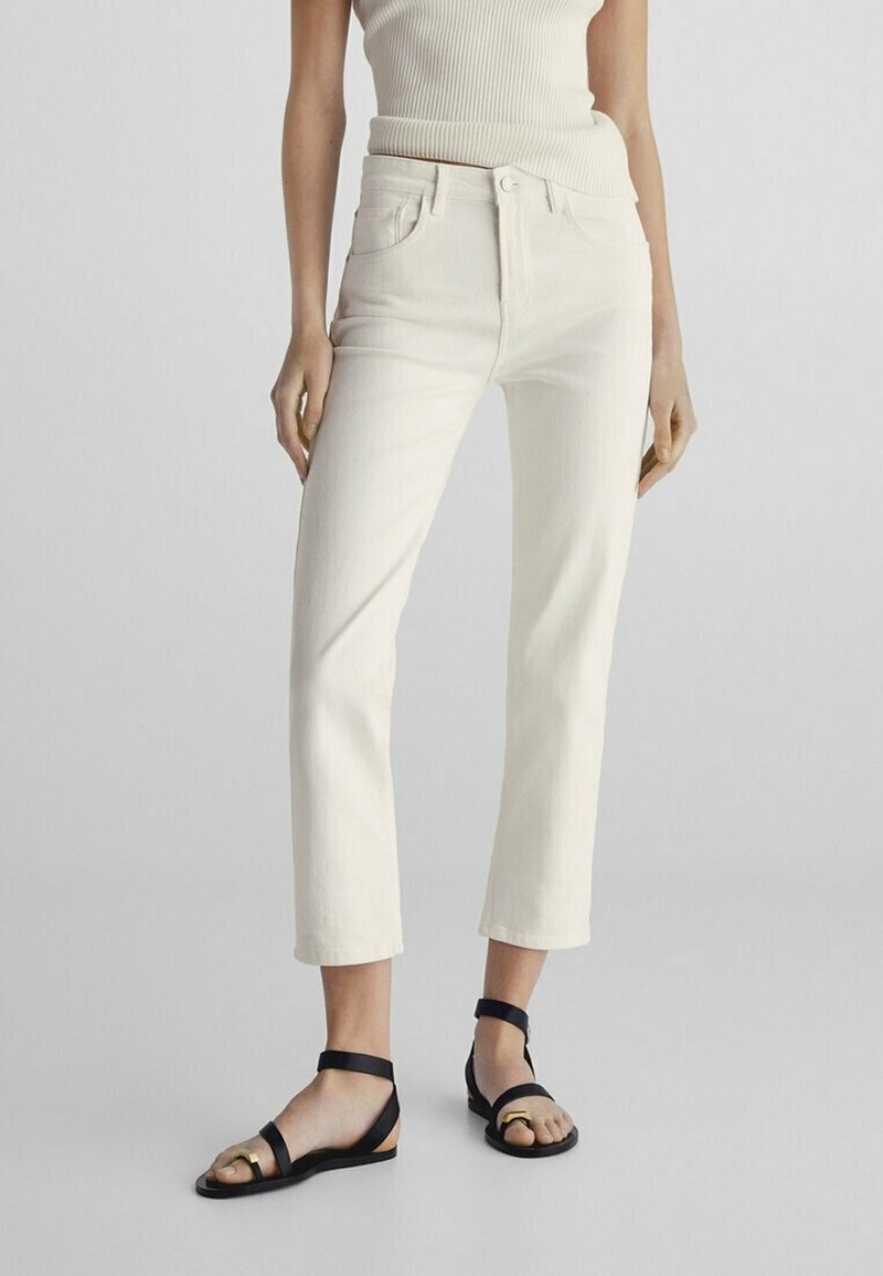 Jean droit blanc avec poches avant, taille haute et texture lisse. Assorti avec des sandales plates noires à brides minimalistes.