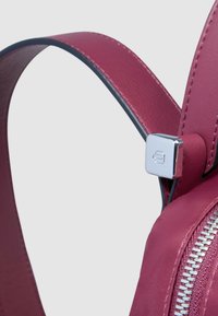 Rucksackriemen aus burgunderrotem Leder mit silbernem Metallelement und schwarzem Rand. Verfügt über eine glatte Textur und eine Reißverschlussdetail an der Seite.