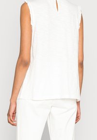 Blusa bianca senza maniche con una finitura testurizzata, caratterizzata da un'apertura a chiave sul retro e bordi sfilacciati sulle maniche.