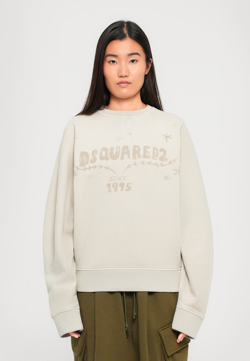 Woman with long dark hair wearing a beige DSQUARED2 sweatshirt and olive green pants, standing against a plain white background.