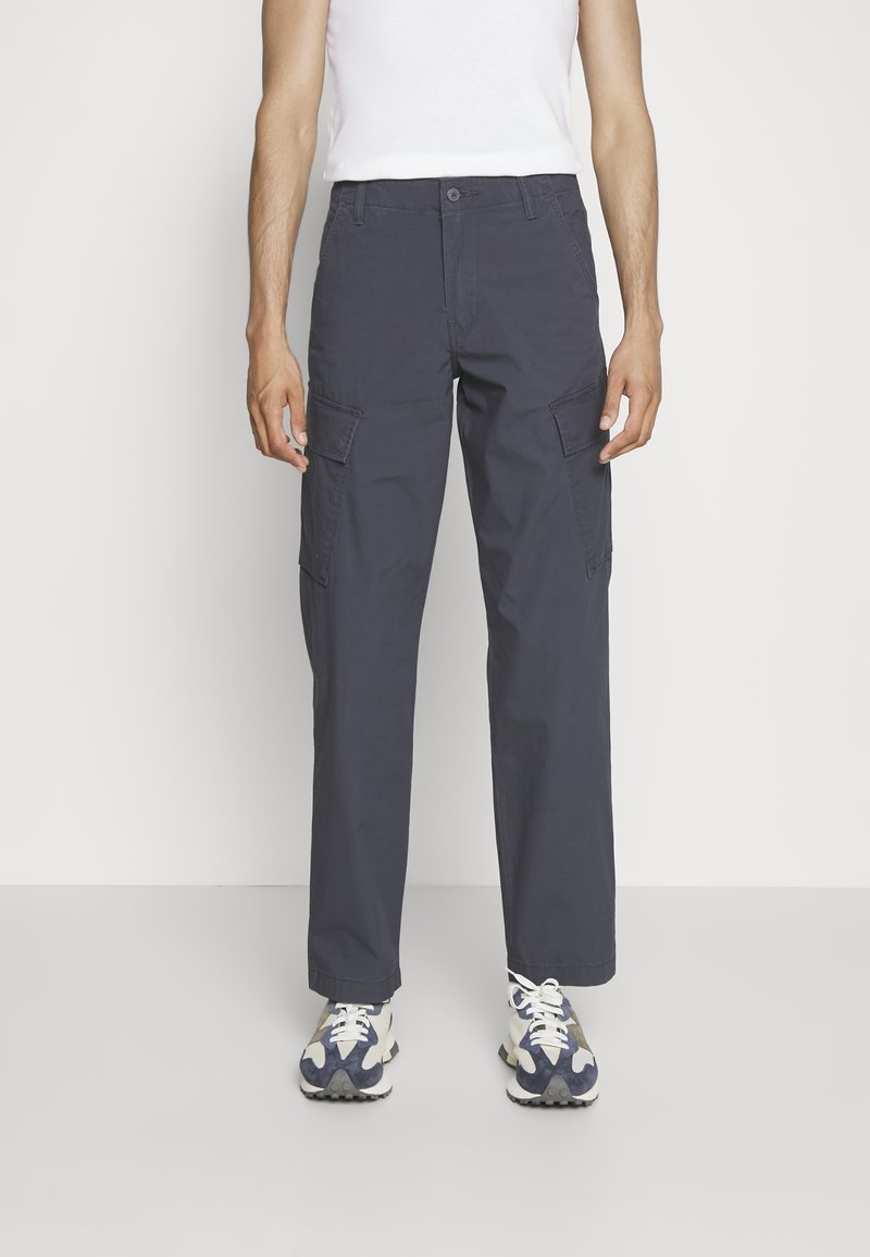 Homme portant un pantalon cargo gris foncé et des baskets blanches, debout devant un fond blanc uni.