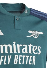 Vihreä urheiluhaalari, jossa on tummansininen kaulus, valkoisella tekstillä "Emirates Fly Better" ja brodeeratulla tykin logolla rinnassa.