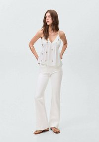 Top blanco sin mangas con dobladillo acampanado y adornos florales dorados, combinado con pantalones acampanados blancos y sandalias marrones. Textura de tela suave.