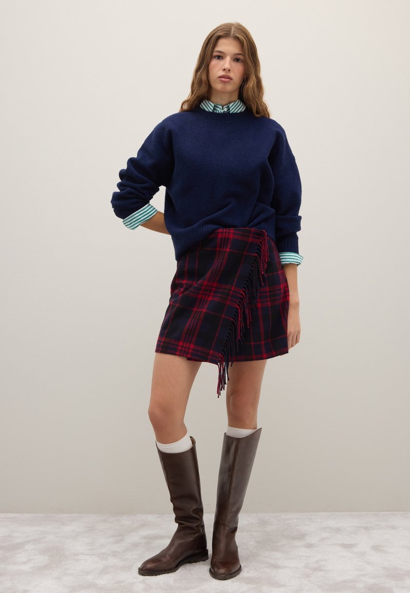 Marineblauer Pullover, karierte Miniröcke mit Fransenrand, braune kniehohe Stiefel und ein gestreiftes Hemd darunter. Glatte Textur und geschichtetes Design.