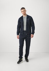 Giacca sportiva blu navy e pantaloni in tessuto leggero, caratterizzati da una chiusura con zip, polsini a costine, vita elastica e dettagli con logo su entrambi i pezzi.