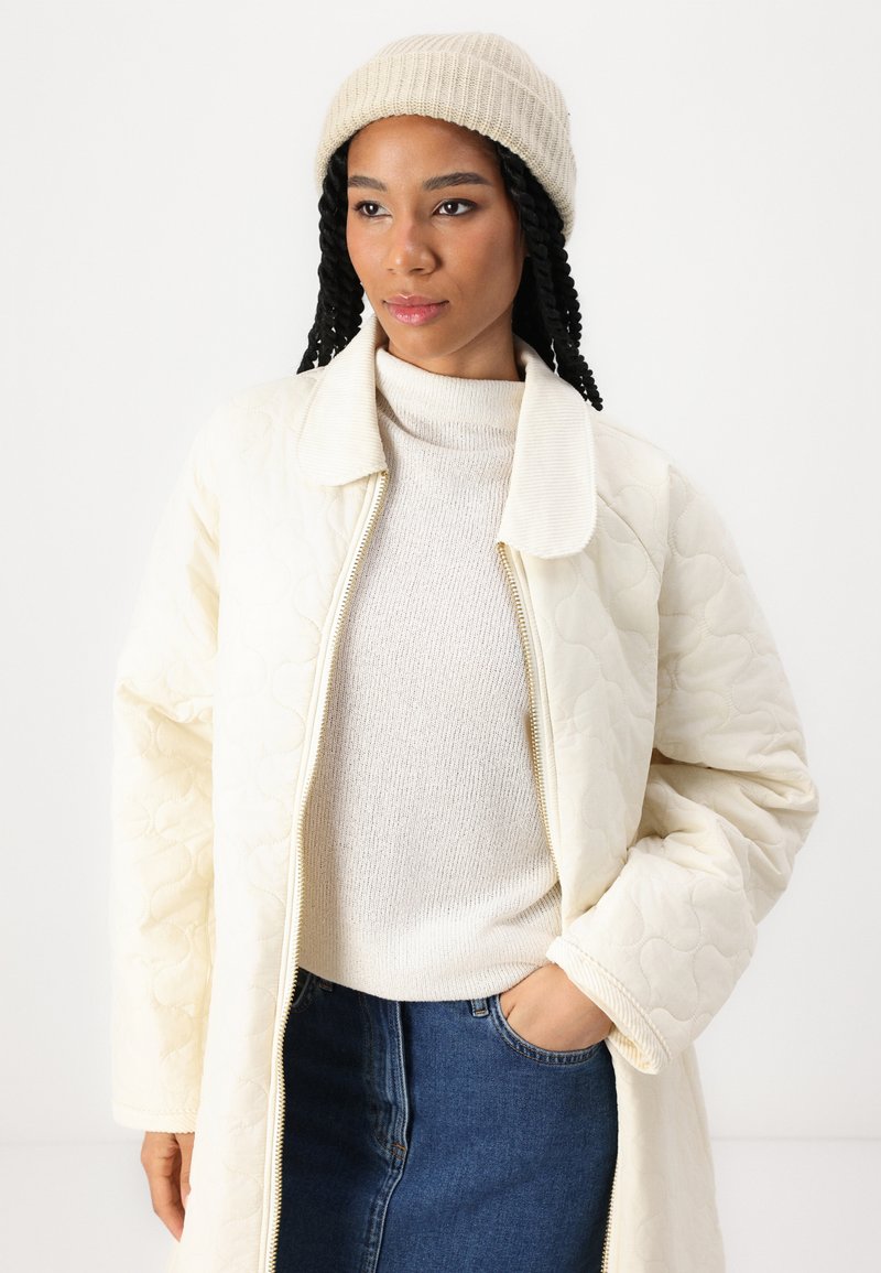 Gep quiltete cremefarbene Jacke mit Kragen, kombiniert mit einem hellbeigen Pullover und einem blauen Jeansrock. Das Model trägt eine gerippte beigen Mütze.