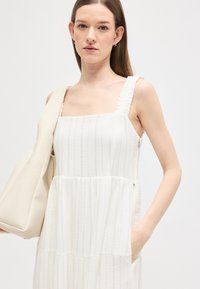 Νεαρή γυναίκα που φοράει λευκό αμάνικο φόρεμα με διακριτικές ρίγες, κρατώντας μπεζ υφασμάτινη τσάντα ώμου, στέκεται μπροστά σε ουδέτερο φόντο.