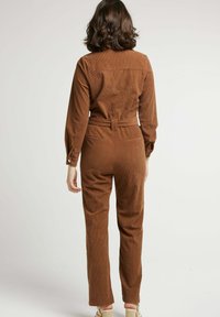 Salopette en velours côtelé marron avec des manches longues, une taille ajustée et des poches arrière. Elle présente une taille ceinturée et un motif texturé en côtes.
