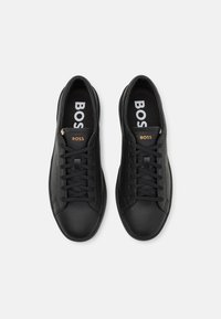 Sneakers in ecopelle nera con suole in gomma, dotate di lacci neri, marchio "BOSS" in bianco sulle solette, e un'etichetta con accento dorato.