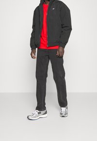 Giacca nera con zip e polsini elastici, t-shirt rossa sotto, pantaloni neri e sneakers argentate lucide con dettagli blu.