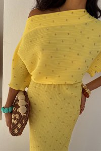 Geel geribbeld jurkje met gouden accenten, met een off-shoulder ontwerp. Een bruine gevlochten clutch en kleurrijke armbanden maken de look compleet.