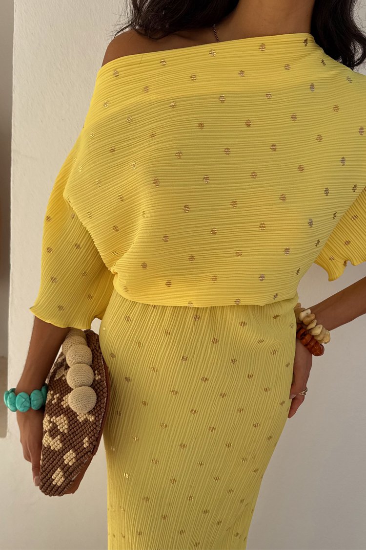 Żółta sukienka prążkowana z złotymi akcentami, z dekoltem typu off-shoulder. Brązowa pleciona torebka i kolorowe bransoletki dopełniają całość.