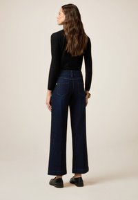 Jeans de mezclilla de tiro alto y pernera ancha en azul oscuro, con dos bolsillos traseros y un acabado suave y texturizado, combinados con una blusa negra de manga larga.
