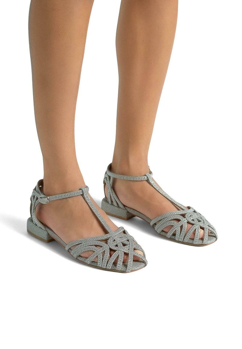 Srebrne, płaskie sandały z paskiem T, z krzyżującymi się wzorami i akcentami w postaci cyrkonii. Otwarta noska, z niskim obcasem.