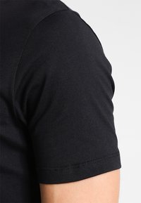 Detailní záběr na horní paži osoby, která má na sobě těsné černé tričko s krátkým rukávem, s viditelným švem na lemu rukávu.