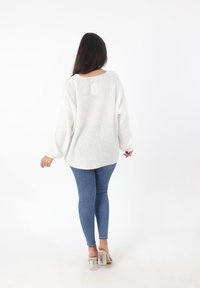 Witte gebreide trui met lange mouwen, textuurpatroon, brede halslijn, gecombineerd met blauwe skinny jeans en transparante sandalen met hoge hakken.