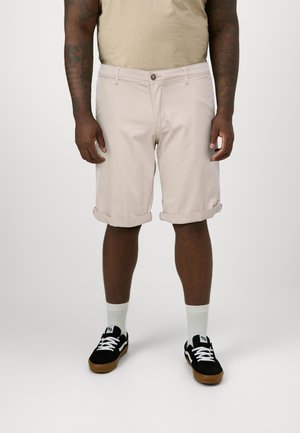 Shorts rose clair avec ourlets roulés, en coton, avec poche avant et détail de bouton marron, associés à des baskets noires et des chaussettes blanches.