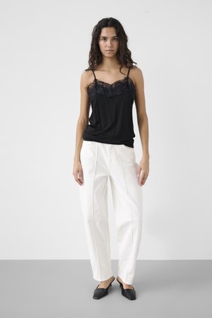 Mujer con cabello largo y rizado, vestida con una camiseta negra con encaje en el borde, pantalones blancos de talle alto y zapatos mule negros de punta estrecha, de pie frente a un fondo liso.