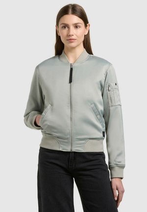 Młoda kobieta w jasnoszarej kurtce bomber z kieszenią na rękawie zapinaną na zamek i czarnych spodniach, stojąca na tle jednolitego jasnego tła.