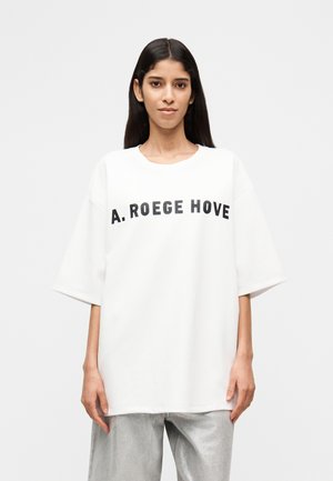 Ženska z dolgimi temnimi lasmi, ki nosi belo široko majico s napisom "A. ROEGE HOVE" in svetle hlače, na enobarvnem ozadju.