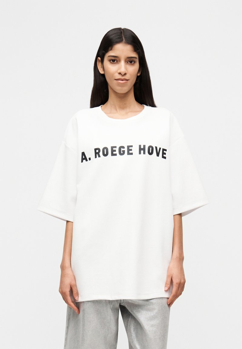 Γυναίκα με μακριά σκούρα μαλλιά που φοράει λευκό φαρδύ t-shirt με το κείμενο "A. ROEGE HOVE" και ανοιχτόχρωμο παντελόνι, σε απλό φόντο.