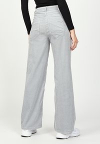 Weite Cordhosen in Hellgrau mit subtilen vertikalen Streifen, hoher Taille und zwei Gesäßtaschen. Die Schuhe sind weiße Sneaker.