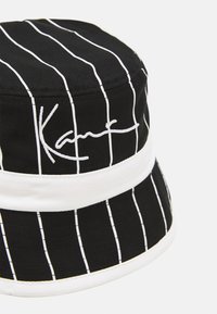 Sort bucket hat med hvide lodrette striber. Har broderet signatur i hvidt og en bred hvid kant rundt om bunden.