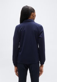 Γυναίκα με μακριά πλεγμένα μαλλιά, που φοράει σκούρο μπλε μπουφάν και αντίστοιχο παντελόνι, στέκεται αντιμετωπίζοντας ένα λευκό φόντο με τα χέρια στις πλευρές.