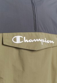 Mesterskabsjakke med en grå overdel og en khaki underdel, prydet med et fremtrædende hvidt logo. Fremstillet af glat, let materiale med lynlås.