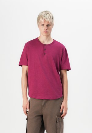 T-shirt tricot à manches courtes de couleur bordeaux avec un col rond et une patte de boutonnage à trois boutons, associé à un pantalon cargo marron ample doté de poches latérales.