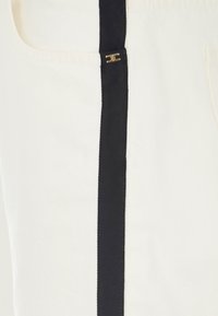 Abito in canvas color crema con una striscia nera verticale. Presenta un dettaglio dorato vicino a una tasca con texture liscia e dettagli di cucitura sottili.