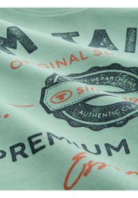 Mintgrünes Baumwoll-T-Shirt mit einem Vintage-Grafikdesign. Mit schwarzem und orangefarbenem Text, einem runden Emblem und einer glatten Textur.
