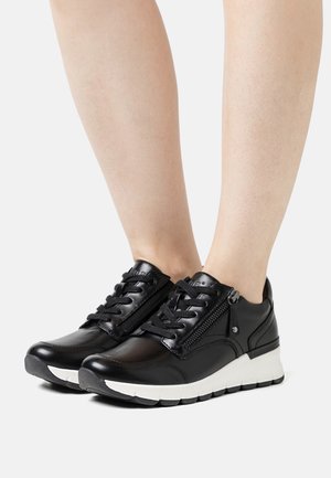 Le sneaker in pelle nera presentano un laccio frontale con un accento in zip, una suola bianca e un design a punta rotonda. Texture liscia e forma sportiva.
