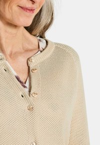 Gilet en tricot beige clair avec un motif texturé, col rond et boutons visibles sur le devant. Matériau doux et respirant.