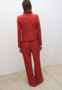 Costum roșu croit care include un blazer ajustat cu rever în colț și pantaloni cu fason evazat. Tesătură netedă, cu un design curat și minimalist.