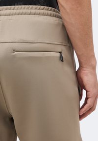 Κοντινό πλάνο μπεζ παντελονιών με ελαστική μέση και μικρή πίσω τσέπη με φερμουάρ, ένα χέρι ακουμπάει δίπλα στο ισχίο.