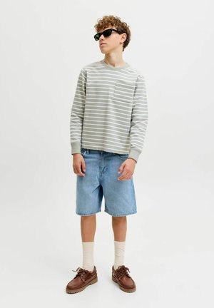 Joven con gafas de sol negras, camiseta de manga larga a rayas blancas y gris claro, pantalones cortos vaqueros, calcetines blancos y zapatos de cuero marrón.