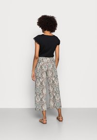 Femme aux cheveux bouclés debout, portant un haut noir à manches courtes, un pantalon large à motifs, et des sandales beiges sur un fond clair uni.