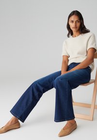 Cremefarbener, fuzzy Strickpullover, dunkelblaue weit geschnittene Jeans und hellbraune Ballerinas, sitzend auf einem Holzstuhl vor einem neutralen Hintergrund.