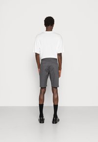 Homme portant une chemise blanche à manches courtes, un short gris jusqu'aux genoux, des chaussettes noires et des chaussures de ville noires, se tenant de dos devant un fond uni.