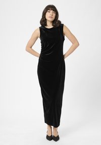 Rochie lungă din catifea neagră, fără mâneci, cu un design drapat și o textură fină. Completată de pantofi negri cu vârf ascuțit, decorați cu ținte albe.